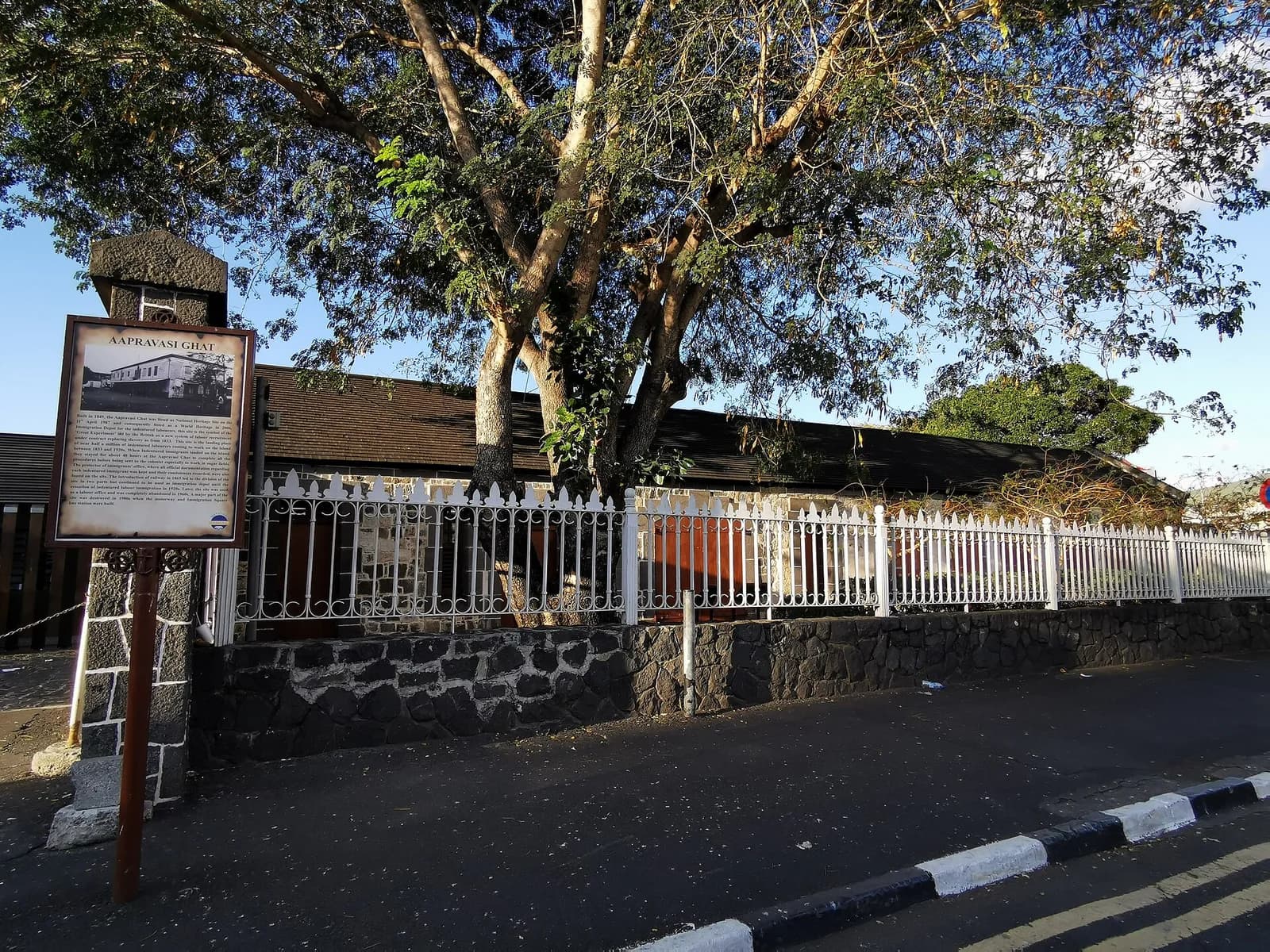 Aapravasi Ghat behind a white fence and large trees, with a heritage information board